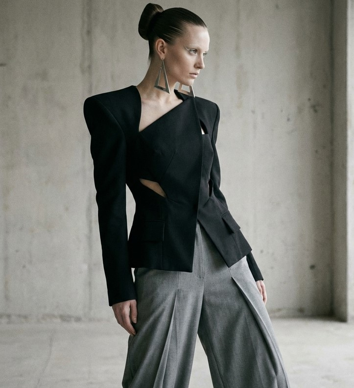 Женщина в асимметричном чёрном жакете со скульптурными плечами и геометрическими вырезами.